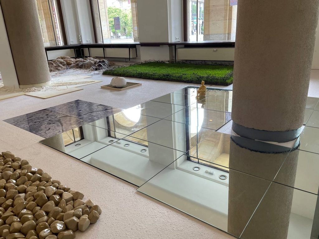 Detail of a mirror installation on the floor, reflecting architectural elements and sculptural objects in the exhibition space.