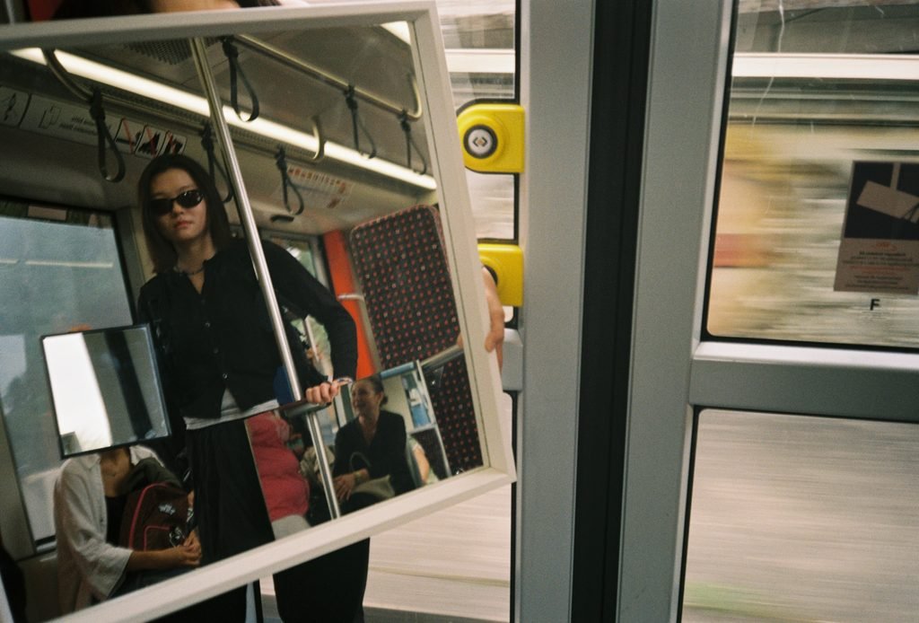 Large mirror inside a moving tram creating layered reflections of the performer, passengers, and interior space.