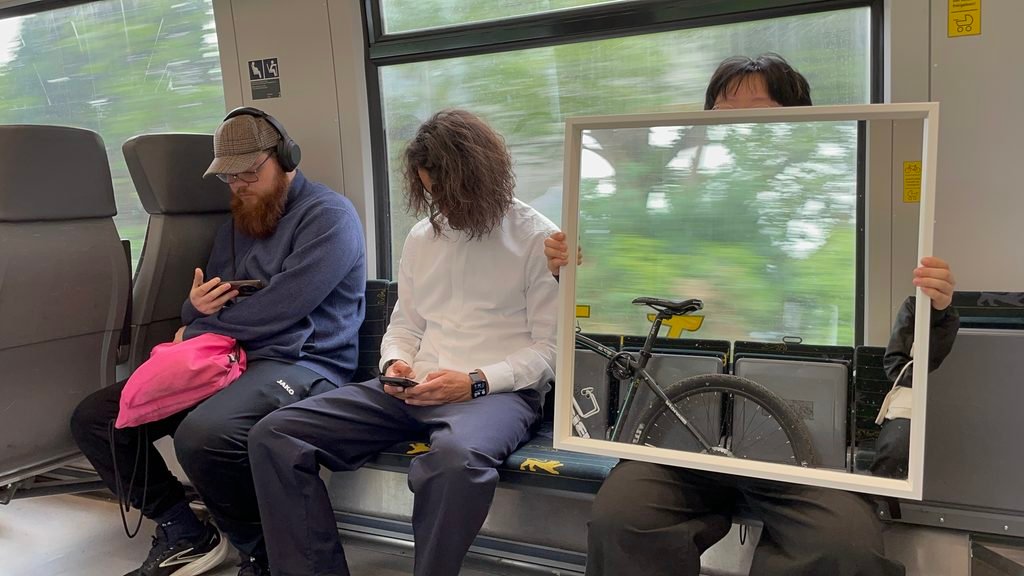 Performer holding a large mirror inside a tram, reflecting the bicycle area and the passing landscape.