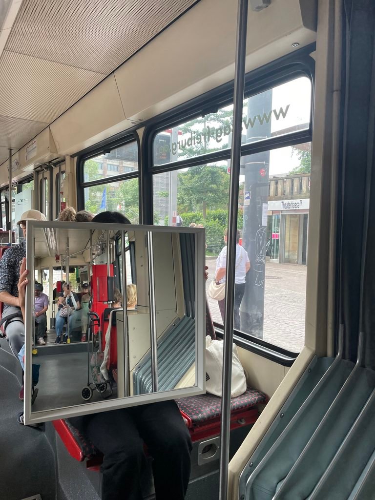 Large mirror in front of a tram window creating a displaced view of the interior and the street outside.