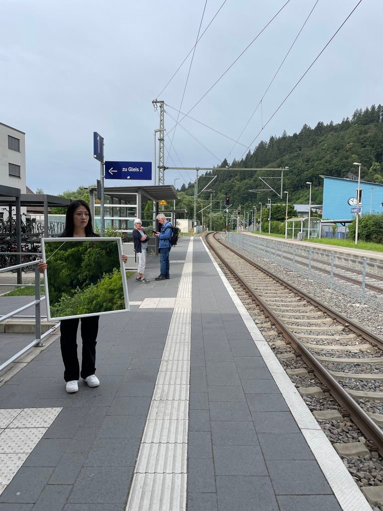Performer holding a large mirror on a tram platform in Freiburg, reflecting trees and the surrounding landscape.